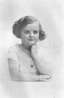 A young girl with light skin tone poses for the camera with one arm in front of her and the other propped up holding her head. Her hair is curled and pinned up. She is wearing a light colored dress with a medallion necklace.