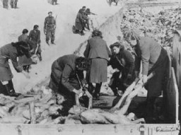 A uniformed soldier stands at the edge of a large burial pit. Groups of light-skinned women in dark clothing carry emaciated bodies by the hands and feet into the mass grave.